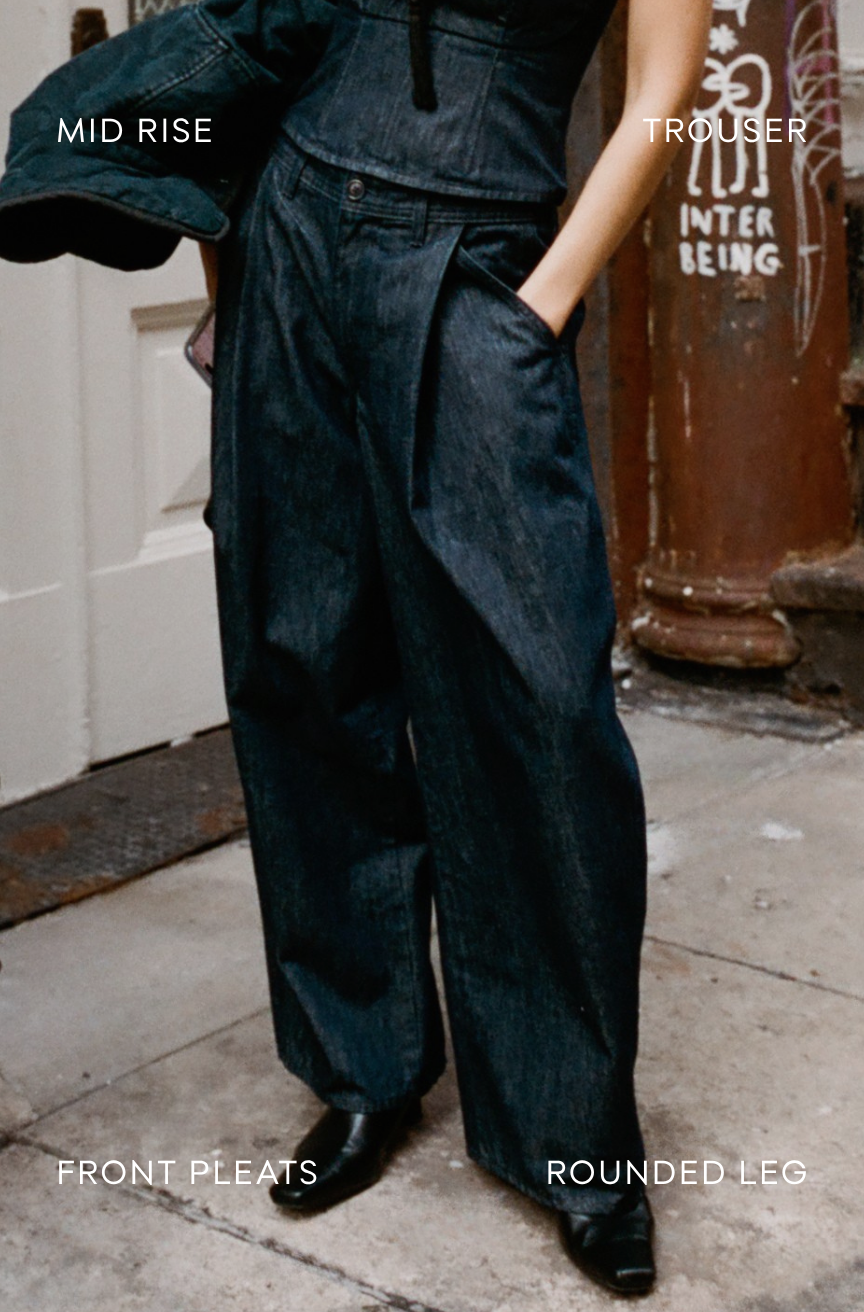A person wearing dark indigo denim wide-leg trousers with front pleats and a cropped matching top, standing on a city sidewalk. White text overlays read 'MID RISE', 'TROUSER', 'FRONT PLEATS', and 'ROUNDED LEG'.